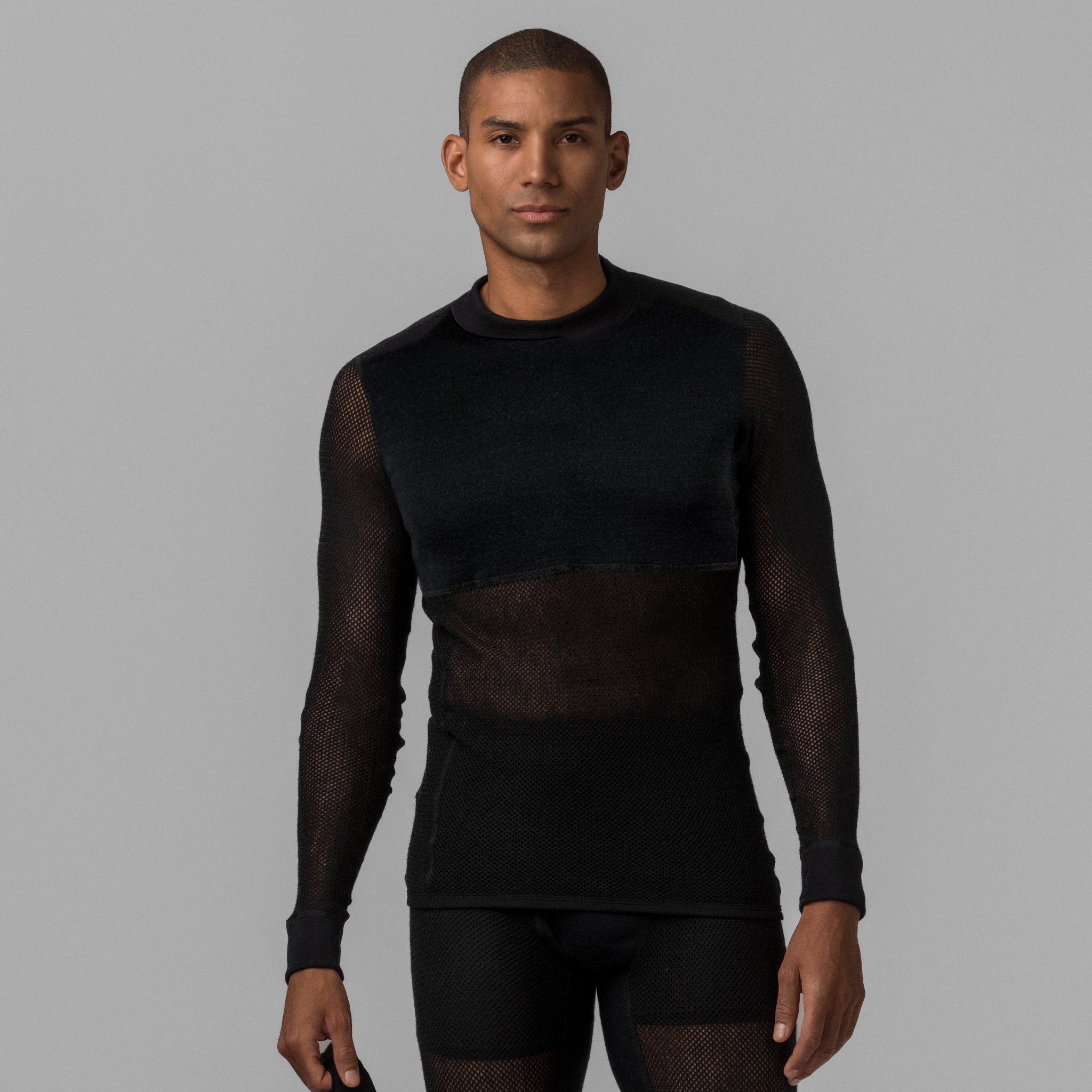 A man faces forward with a neutral expression, wearing the Janus Wool Jersey mesh merino - Black Wool 165 Unisex top and matching bottoms against a plain gray background, showcasing this stylish unisex look.