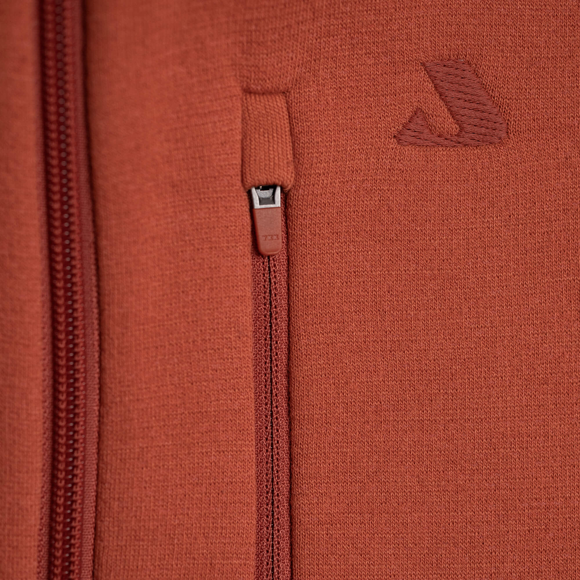 Close-up of the Jacket Merino – AirWool 350 Men in rust, featuring a visible merino wool texture, zipper, and a small embroidered logo above the pocket with the zipper pull centered.