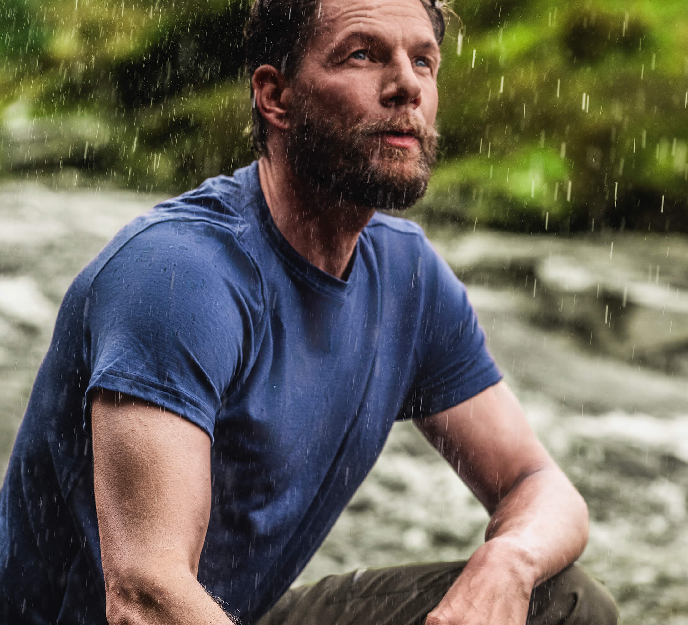A bearded man in a wet blue merino t-shirt sits outdoors in the rain, looking up thoughtfully. Blurred greenery and water are visible in the background.