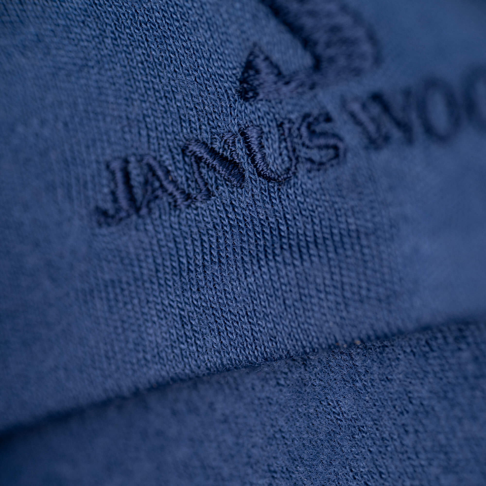 Close-up of blue fabric with partially visible "WOOL" embroidery and triangular logo. The texture showcases the warmth and breathability typical of the Beanie Merino – Airwool 260.
