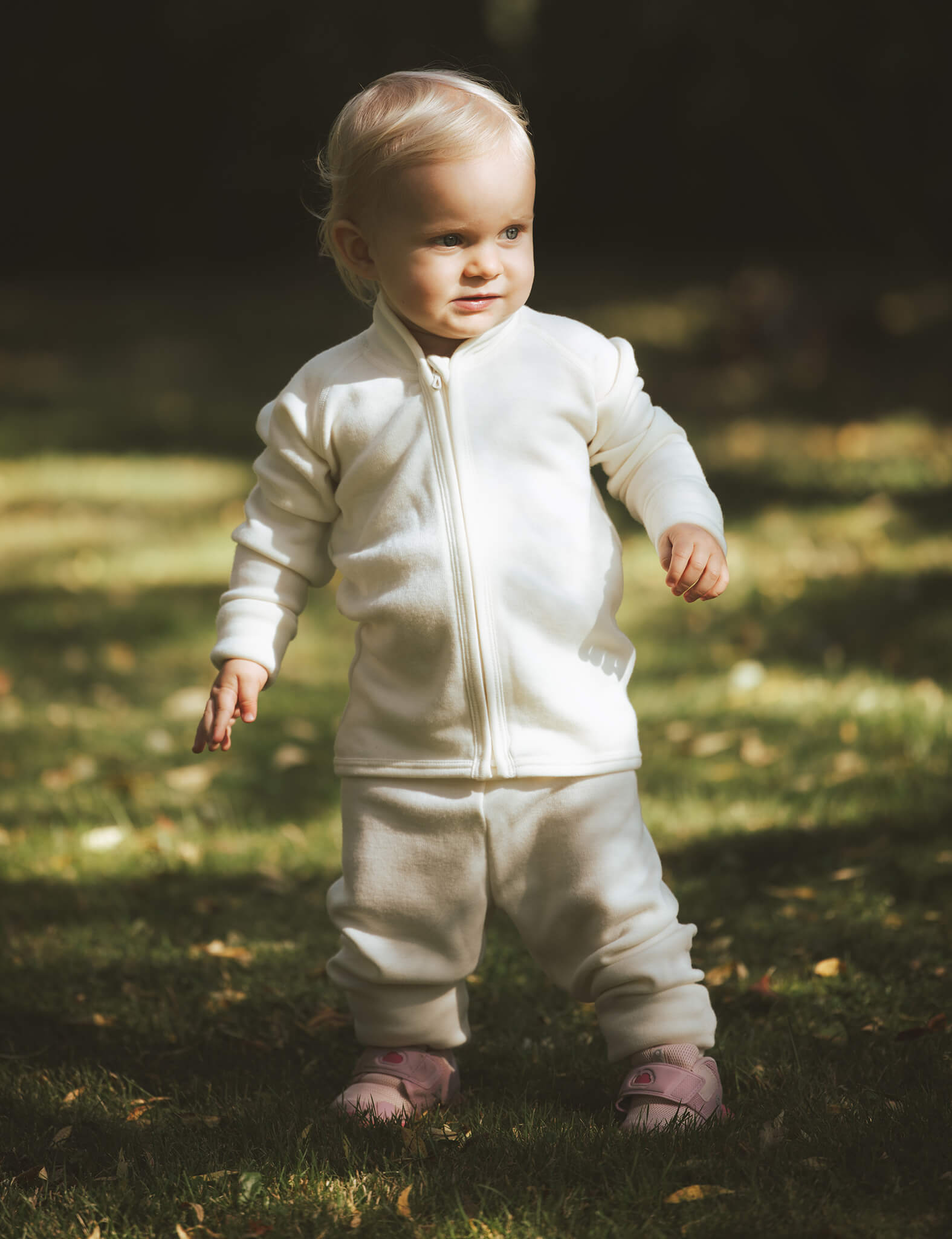 A toddler with blonde hair stands on grass in sunlight, wearing a Marshmallow-colored Merino Baby AirWool zip jacket and pink shoes, looking slightly to the side.