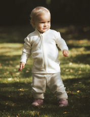 A toddler with blonde hair stands on grass in sunlight, wearing a Marshmallow-colored Merino Baby AirWool zip jacket and pink shoes, looking slightly to the side.