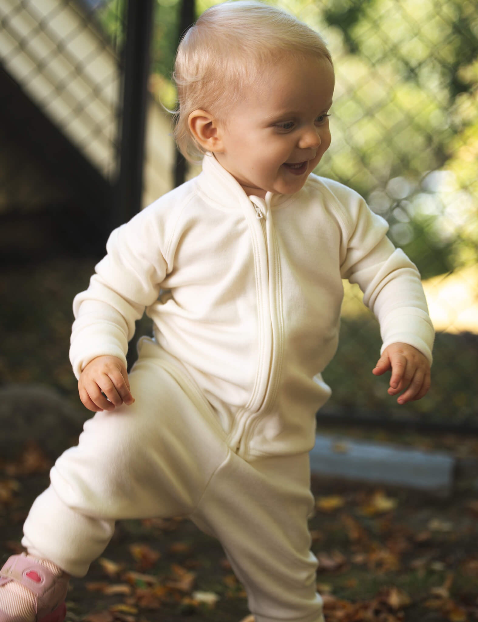 A smiling toddler stands outdoors on leafy ground, lifting one foot, wearing the Merino Baby AirWool jacket in cream, with a fence and greenery in the background.
