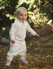 A smiling toddler with blonde hair, dressed in a white outfit and pink shoes, stands outside on grass and autumn leaves with a blurred background of trees, wearing the Jacket – Merino Baby AirWool.