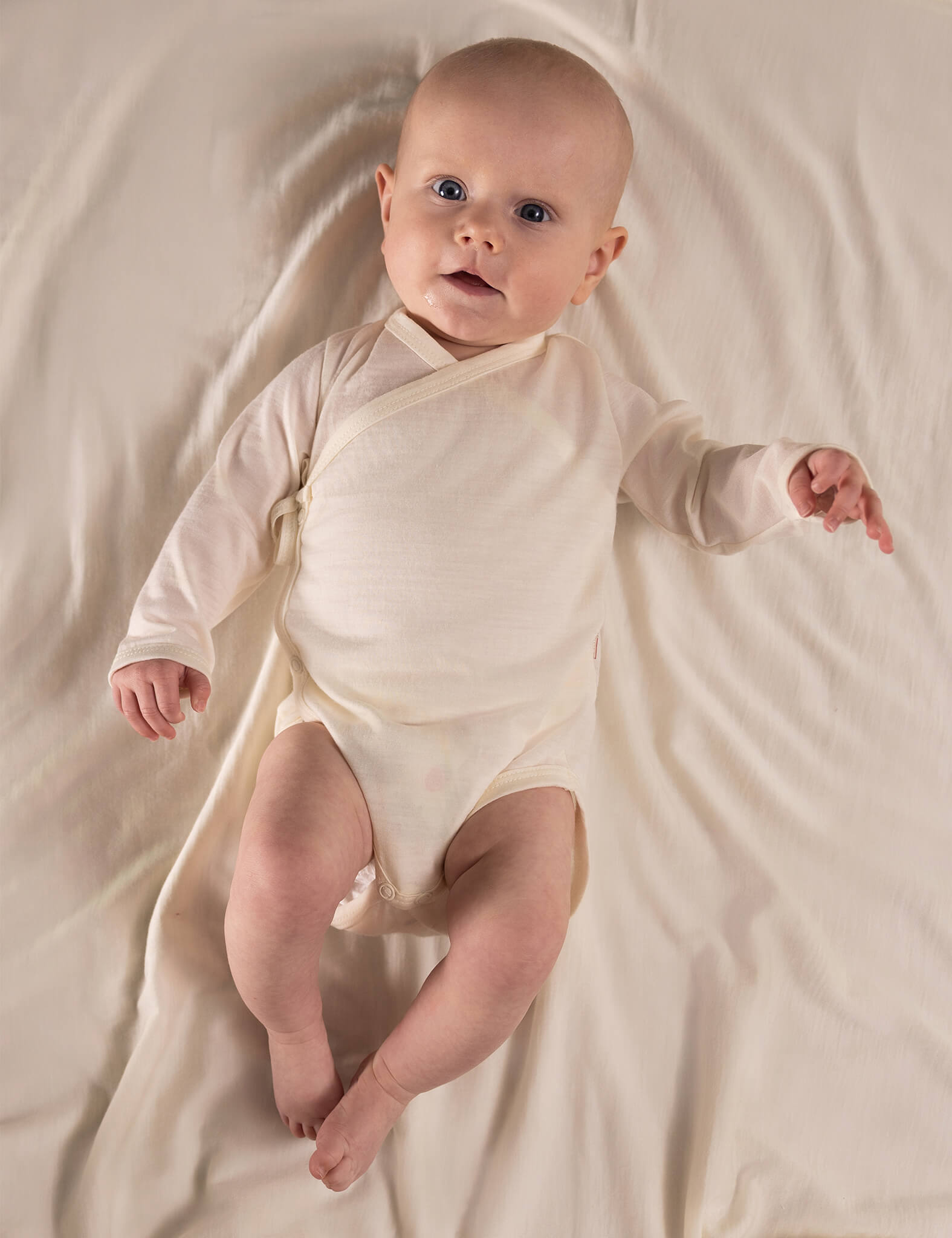 A baby wearing the Wrapover Bodysuit – Merino Baby Light in Marshmallow lies on a soft, matching blanket, looking up with bright eyes and one arm slightly raised.
