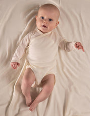 A baby wearing the Wrapover Bodysuit – Merino Baby Light in Marshmallow lies on a soft, matching blanket, looking up with bright eyes and one arm slightly raised.