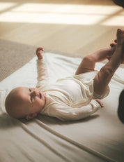 A baby wearing the Wrapover Bodysuit – Merino Baby Light in a cream color lies on a soft blanket, smiling up as someone gently holds their legs. Warm sunlight streams in from the left, highlighting the breathable fabric.
