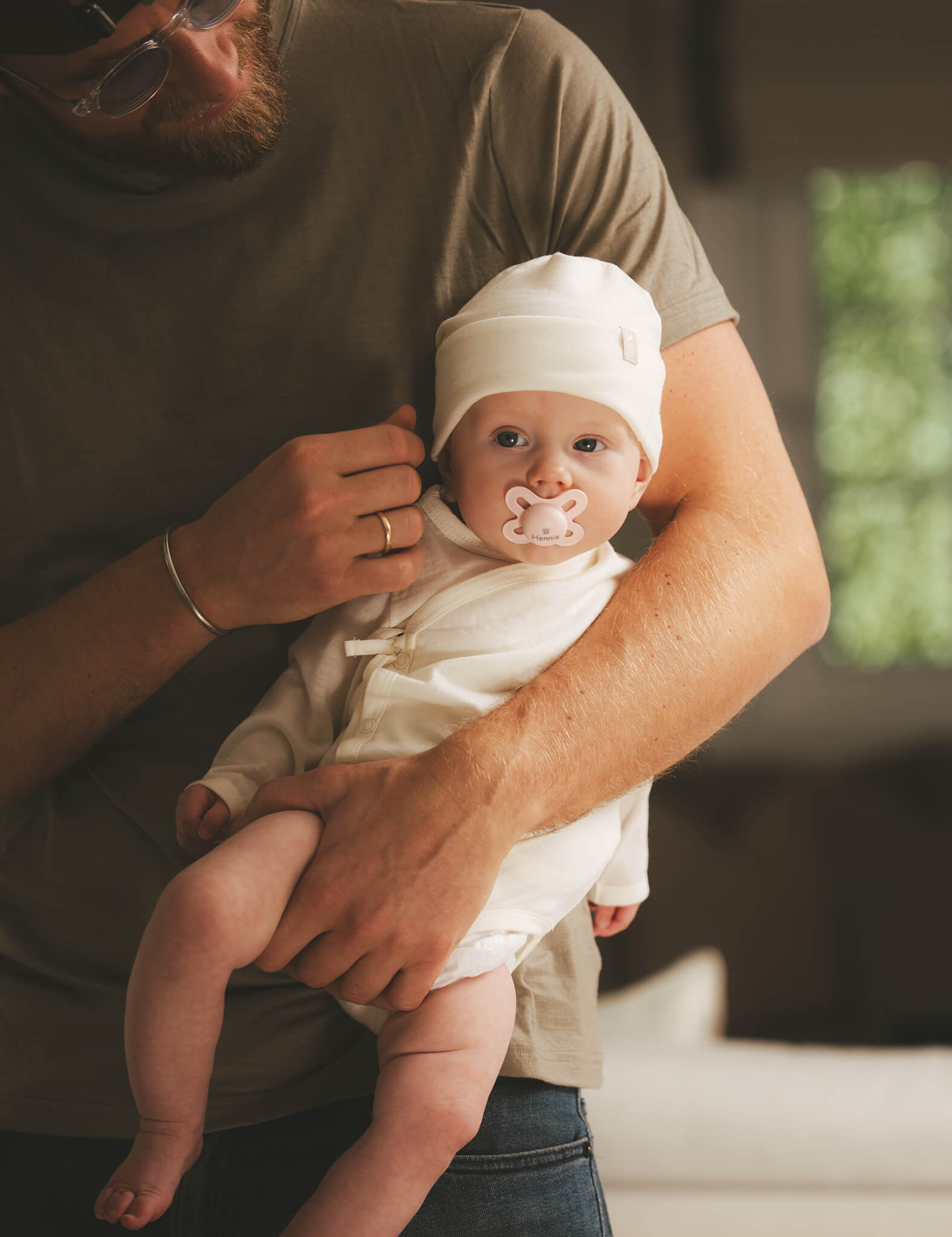 A man gently holds a baby dressed in the Wrapover Bodysuit – Merino Baby Light, complete with a pacifier and hat. The baby looks at the camera while sitting in the man’s arms as warm, soft lighting fills the scene.