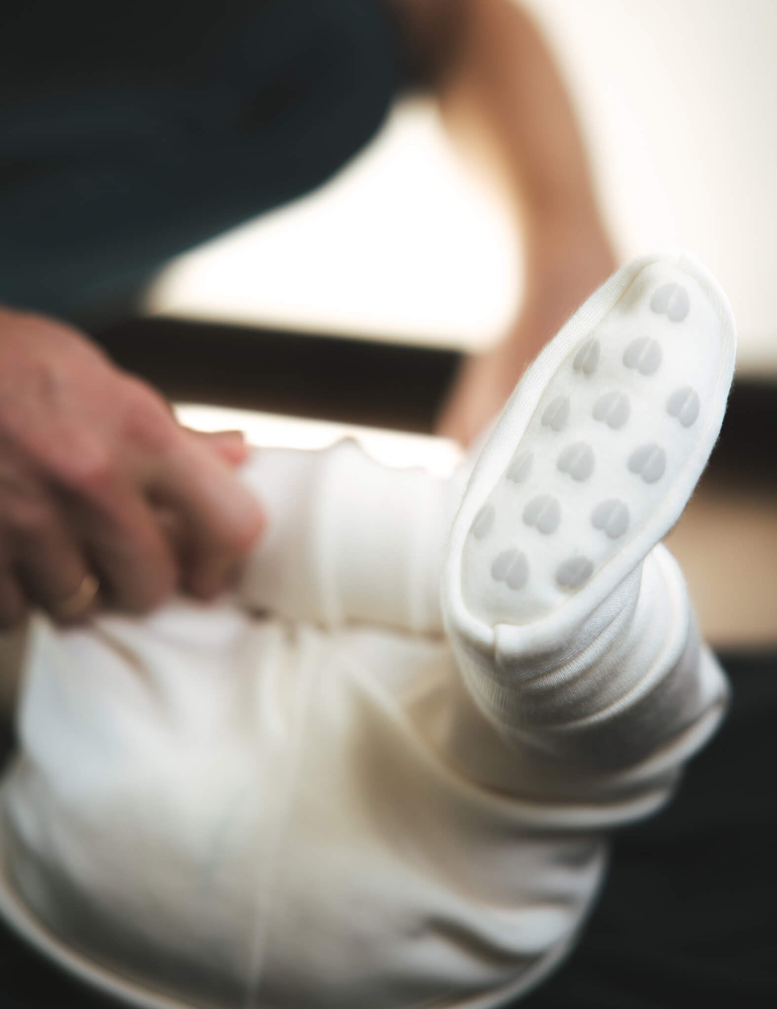 A close-up of an adult holding a baby dressed in white, focusing on the baby’s foot in warm, breathable Booties – Merino Baby AirWool in the color Marshmallow. The background is softly blurred.