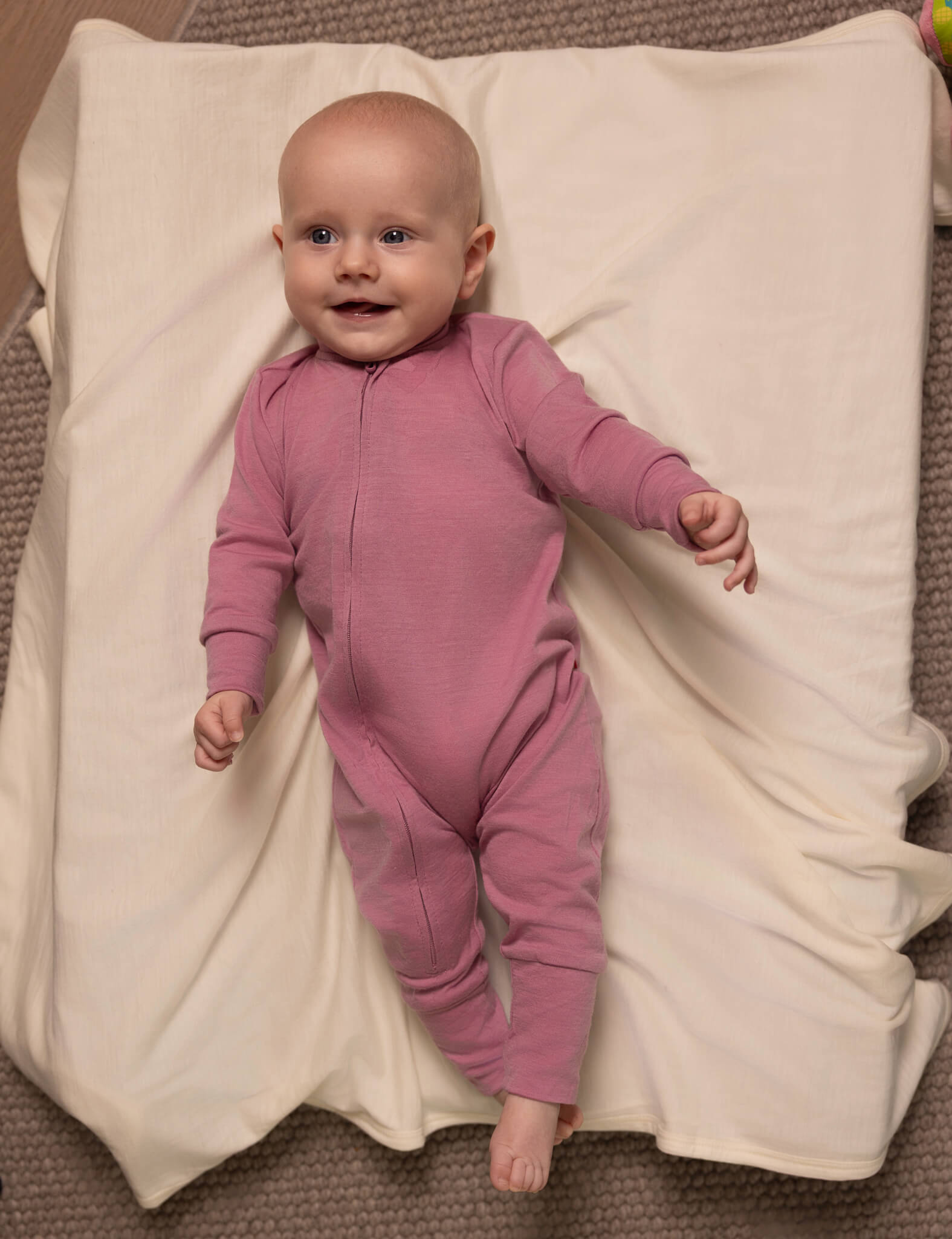 A smiling baby in a pink onesie lies on the cream-colored Blanket – Merino Baby AirWool atop a textured surface, looking up with one arm bent and one hand slightly open.