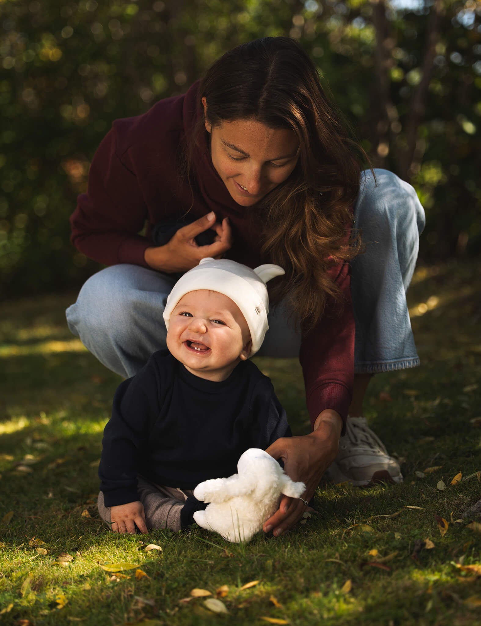 A woman crouches behind a smiling baby in a white hat and dark shirt, who is sitting on the grass holding a white stuffed toy. Sunlight filters through trees in the background, creating a perfect setting for enjoying the cozy comfort of the Pullover – Merino Baby AirWool.