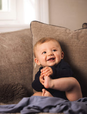A smiling baby with light hair sits on a brown couch, wearing the Body – Merino Baby AirWool bodysuit and holding their foot with both hands. A window and grayish blanket are visible in the background.