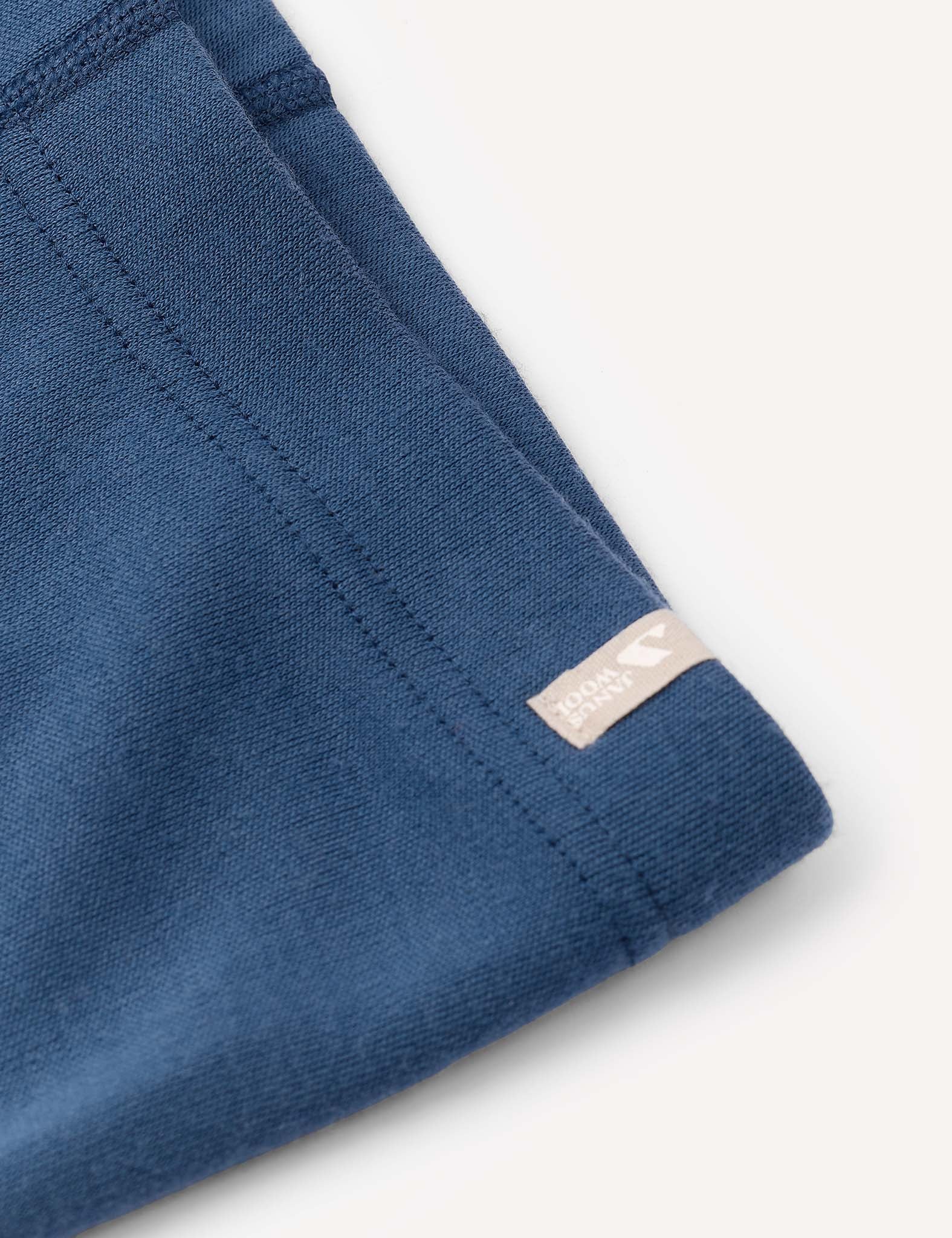 Close-up of a blue fabric garment—the Leggings – Merino Kids AirWool—crafted from brushed AirWool, featuring a stitched seam and a small beige tag with white text and logo, set against a plain light background.