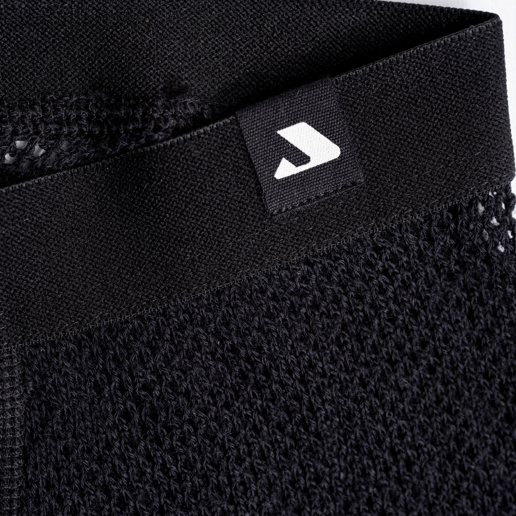 Close-up of the black, textured knit waistband on Leggings mesh - Norse Merino 165 Unisex. A sewn-on black tag with a white geometric logo is visible, highlighting the stitching, fabric details, and wool mesh texture.