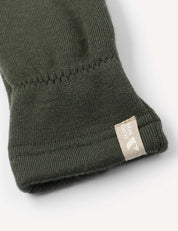 Close-up of the cuff of a dark green Merino Kids AirWool mitten, featuring a small beige fabric tag sewn onto the edge with a white logo and text.