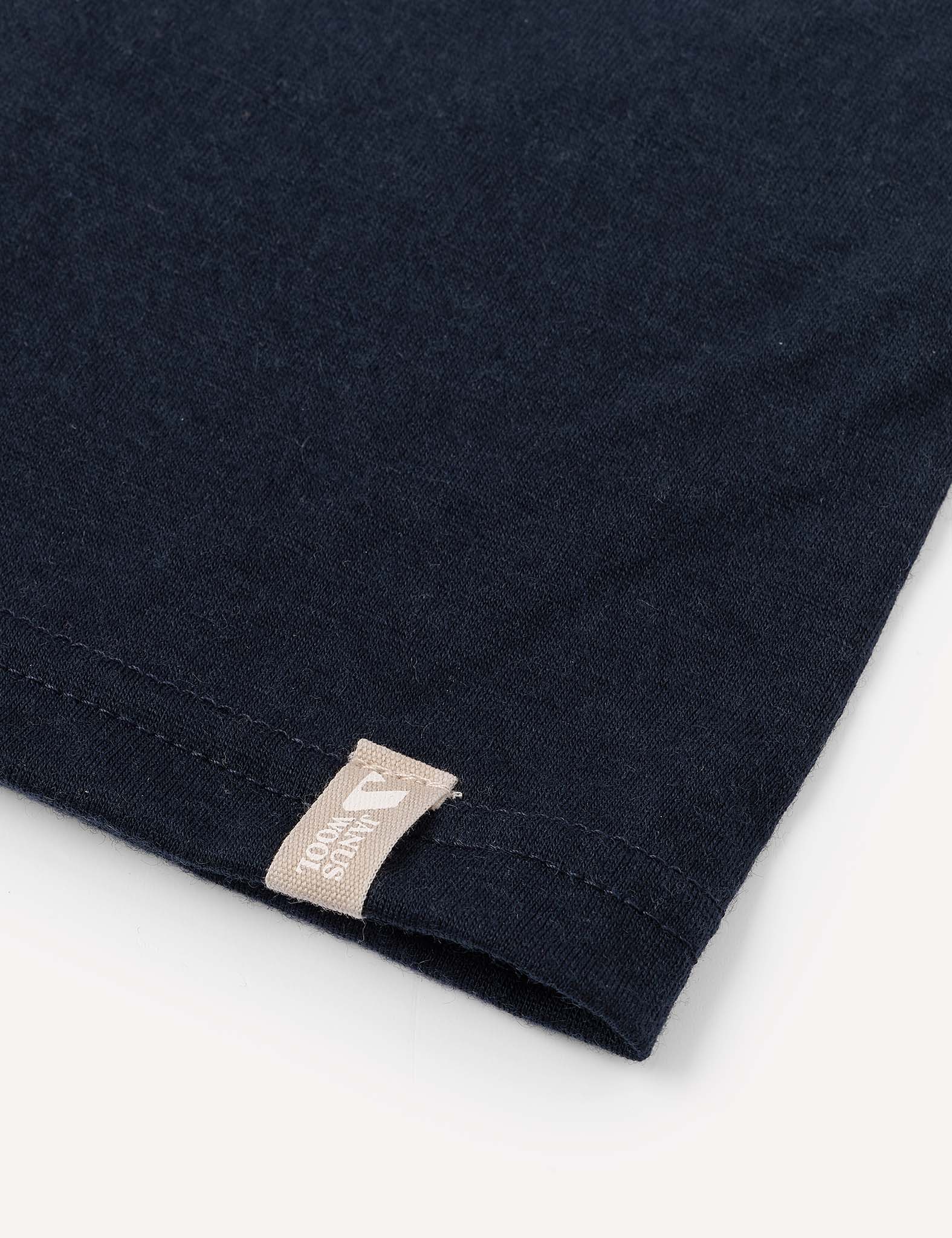 Close-up of the hem of the dark-colored Neckwarmer – Merino Kids Light, crafted from merino wool fabric, with visible stitching. A small beige tag featuring text and a graphic is sewn into the seam near the edge. The background is a plain, light surface.