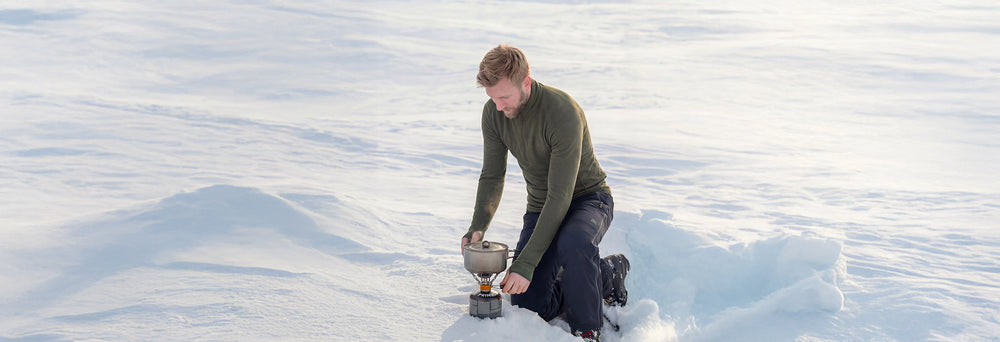 Person in Janus Wool double layer zip polo and black pants standing in a snowy landscape, using a gas stove on a snowdrift.