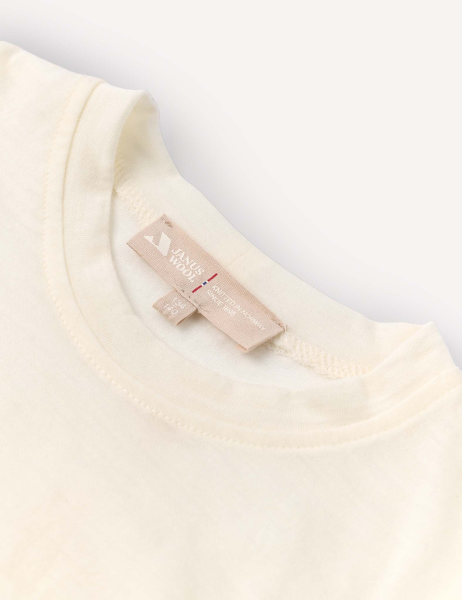 Close-up of the neckline of the T-Shirt – Merino Kids Light in a cream shade, featuring a beige clothing tag with printed text and symbols, stitched inside the collar. Renowned for its natural temperature regulation and all-day comfort, the merino wool shirt is displayed flat on a light background.