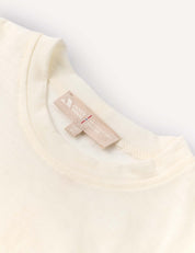 Close-up of the neckline of the T-Shirt – Merino Kids Light in a cream shade, featuring a beige clothing tag with printed text and symbols, stitched inside the collar. Renowned for its natural temperature regulation and all-day comfort, the merino wool shirt is displayed flat on a light background.