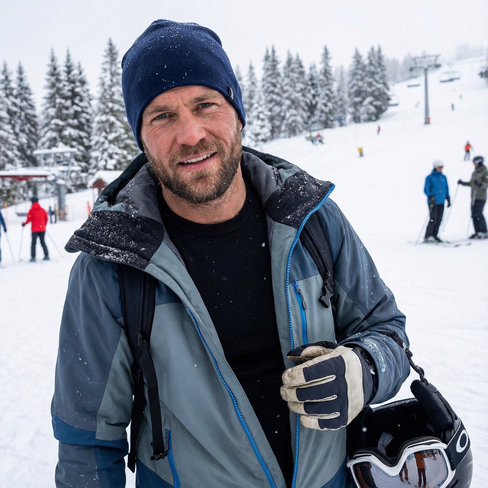 A man wearing a Beanie merino - Classic 260, winter clothes, and gloves stands on a snowy ski slope holding ski goggles, with snow-covered trees and skiers in the background.