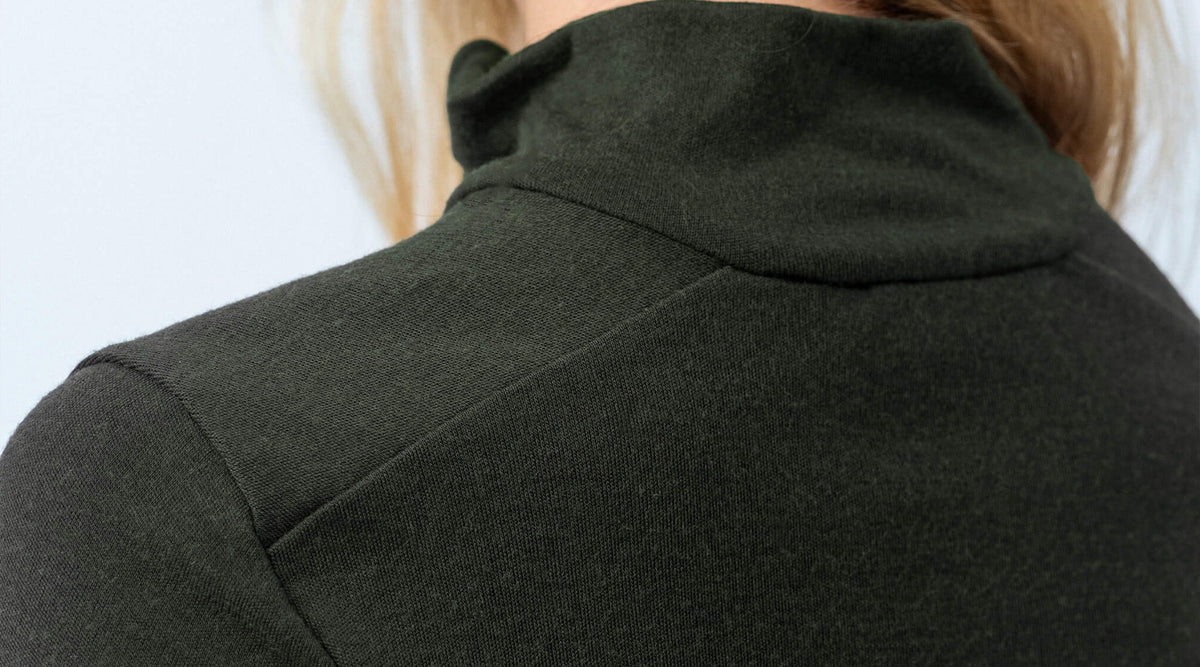 Close-up of the back of a person wearing a dark green merino wool turtleneck sweater, with light brown hair partially visible and a neutral light background.